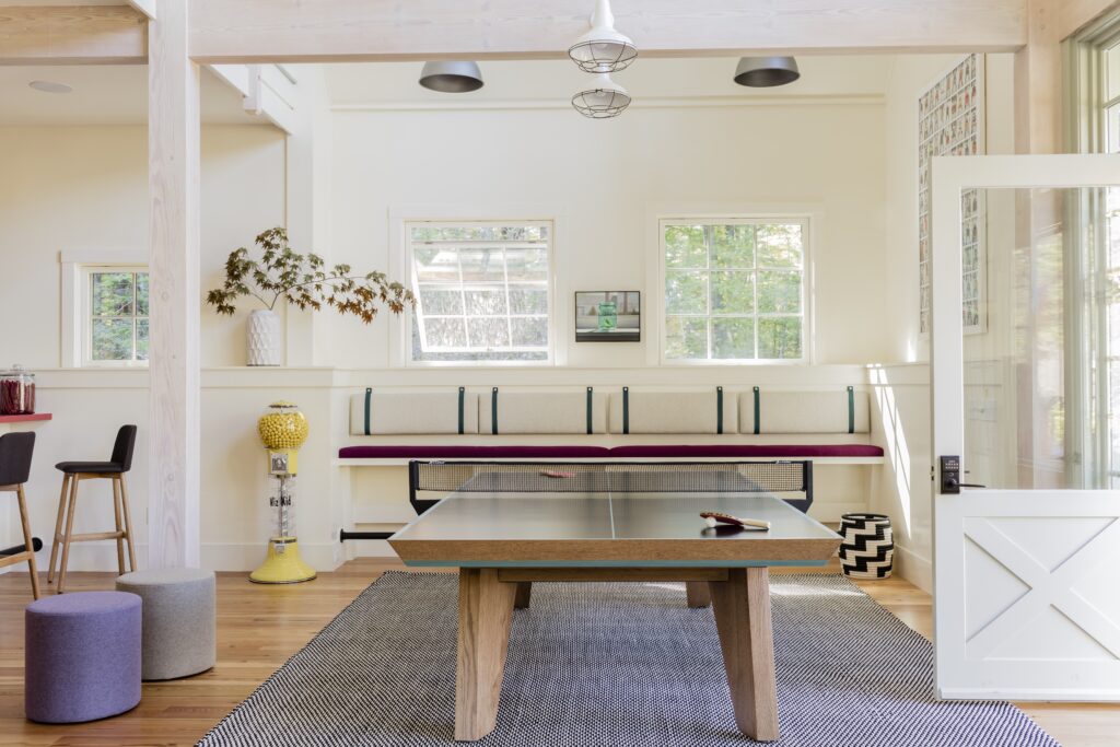 A bright, modern game room featuring a Recent Work - 11 Ravens ping pong table at its center, a cushioned bench under three windows, a zigzag-patterned stool, bar stools, and a yellow gumball machine on a wooden floor.